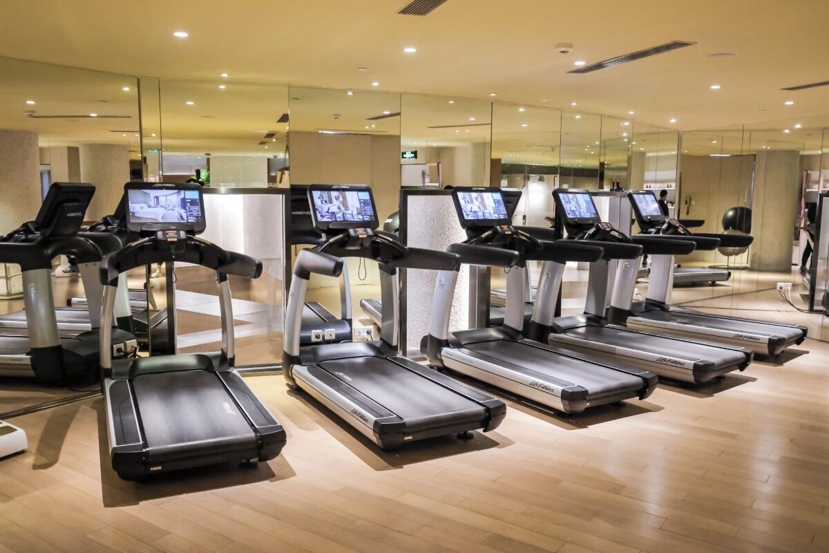 A gym with multiple treadmills, each equipped with a digital screen, in a modern indoor setting with wooden flooring and mirrored walls.