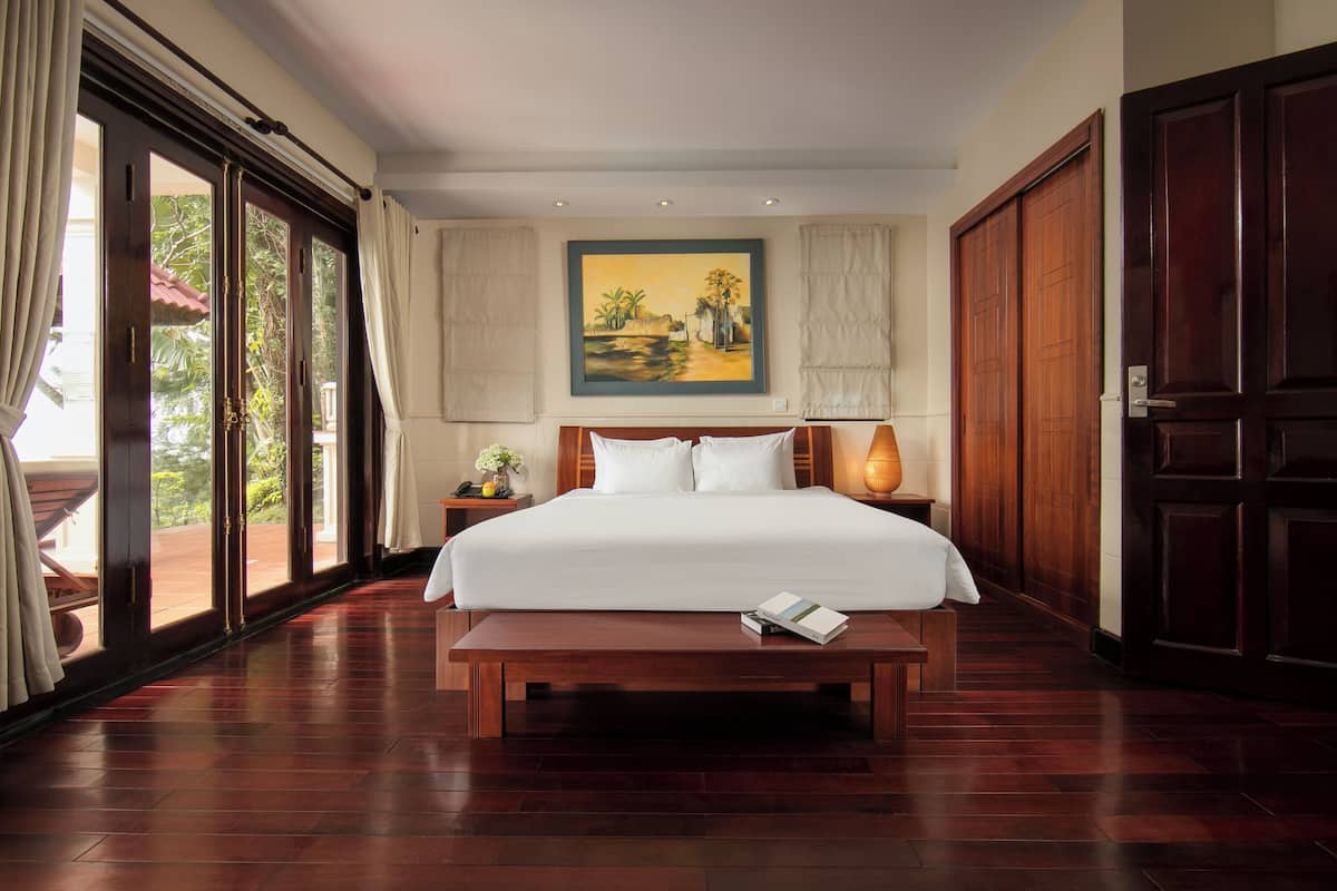 A hotel room with a large bed, a wooden bench, a painting on the wall, and a view through the glass doors.
