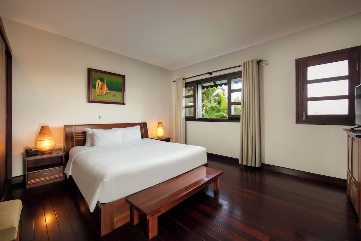 A hotel room with a large bed, wooden bench, two bedside lamps, a painting on the wall, a window with curtains, and a view of greenery outside.
