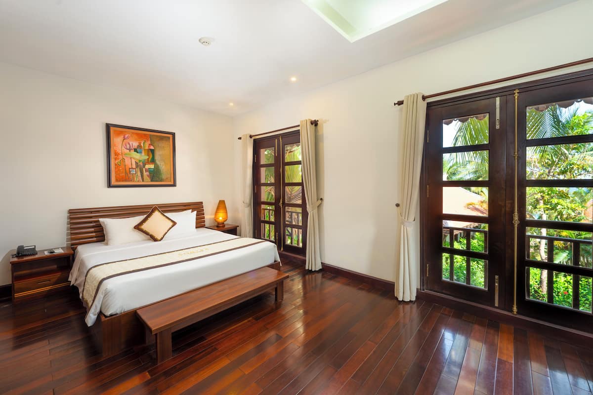 A spacious bedroom with a large bed, wooden flooring, a nightstand, a bedside table, and a window with curtains.