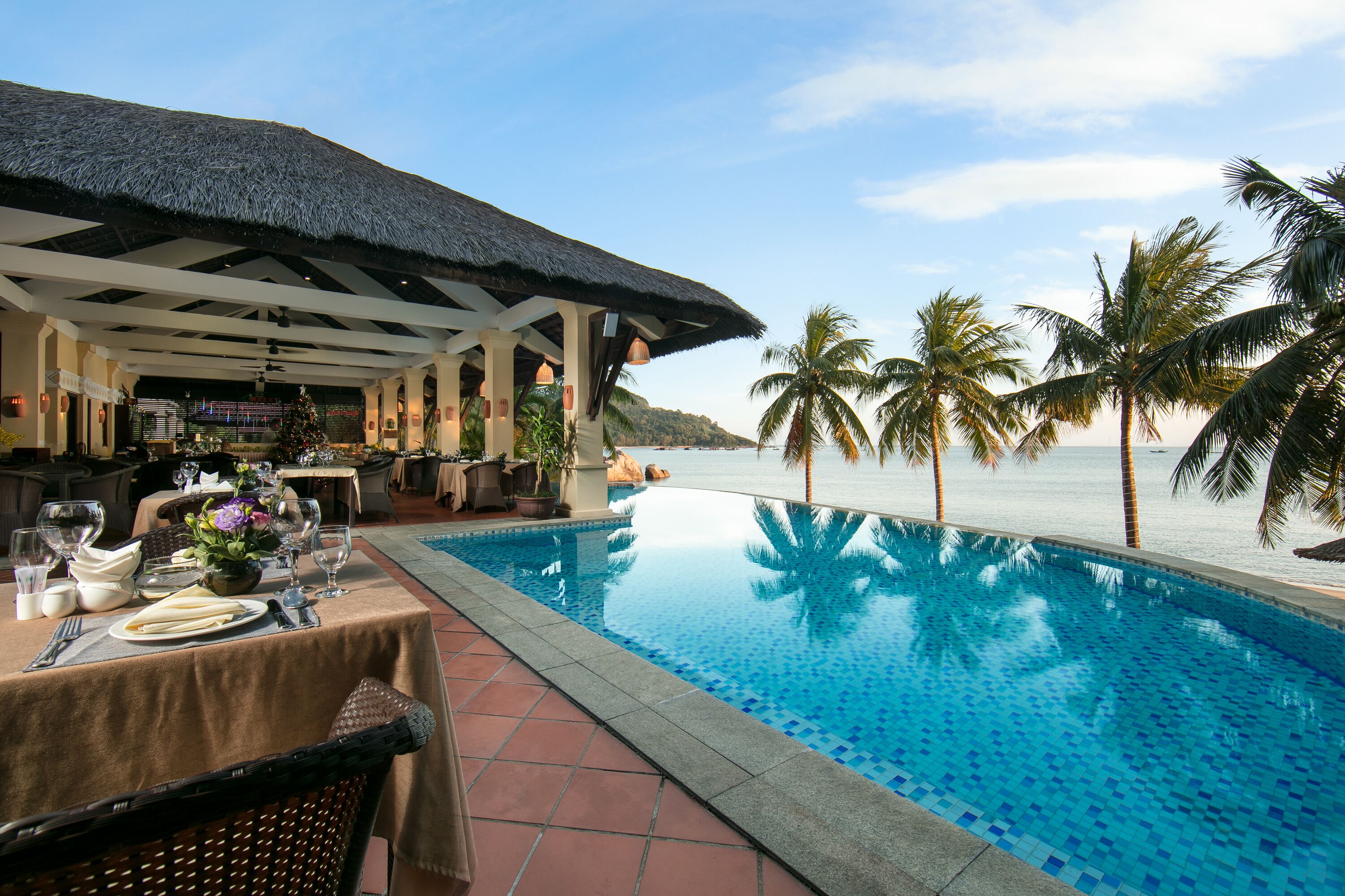 A poolside dining area with a table set for two, overlooking a clear blue pool and a beach.