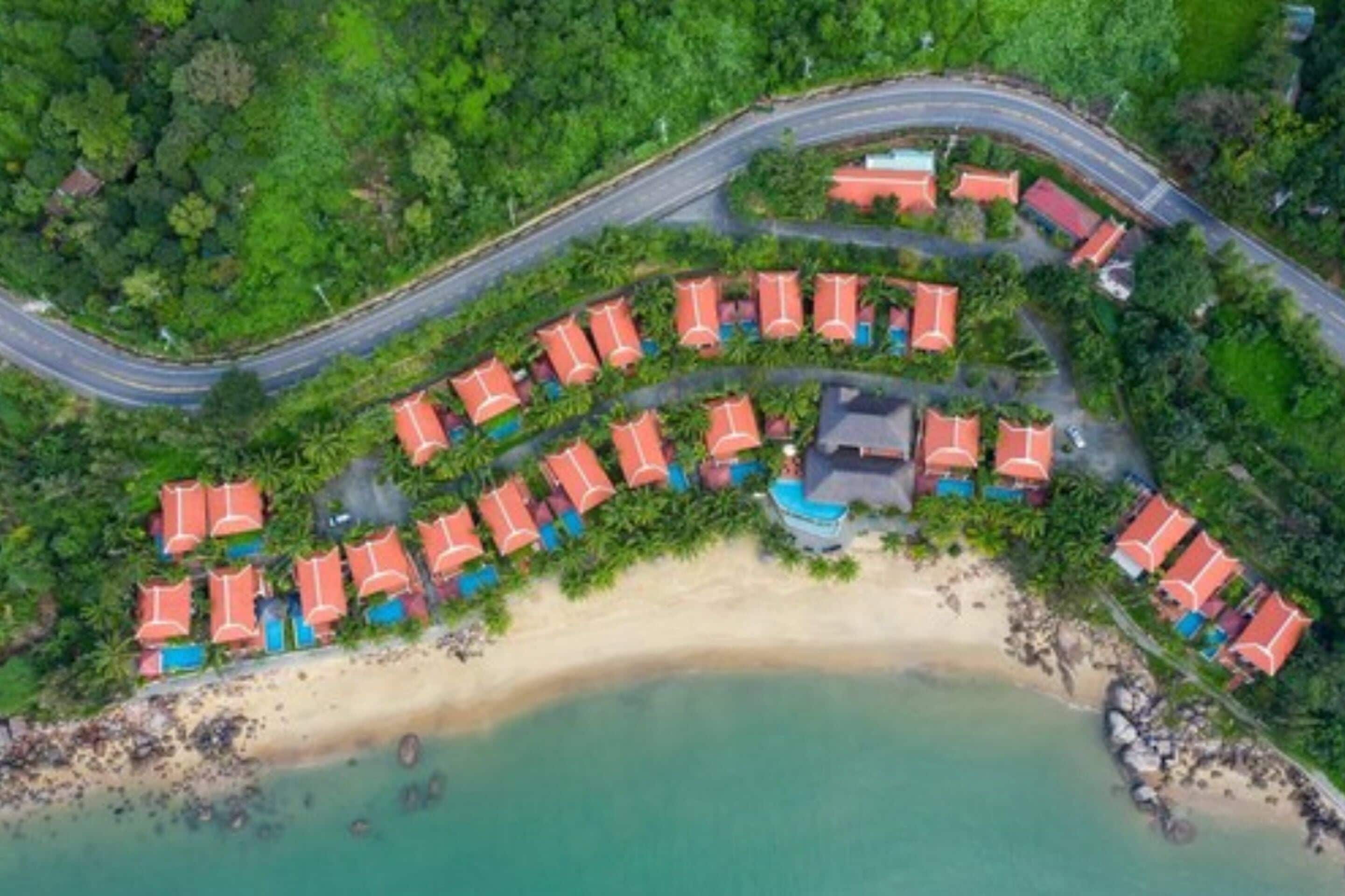 An aerial view of a resort with a curved road, beach, and bungalow-style accommodations.