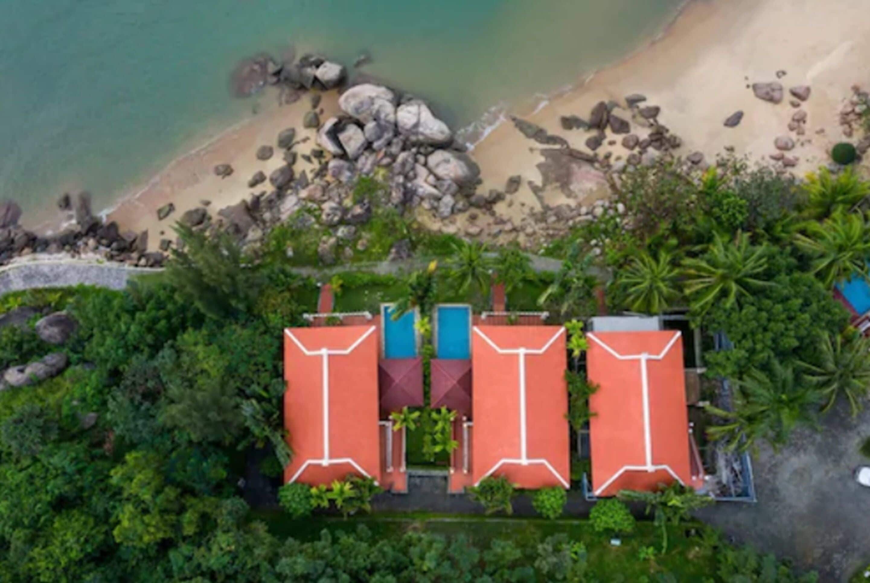 An aerial view of a resort with three red-roofed buildings, a sandy beach with rocks, and lush greenery.