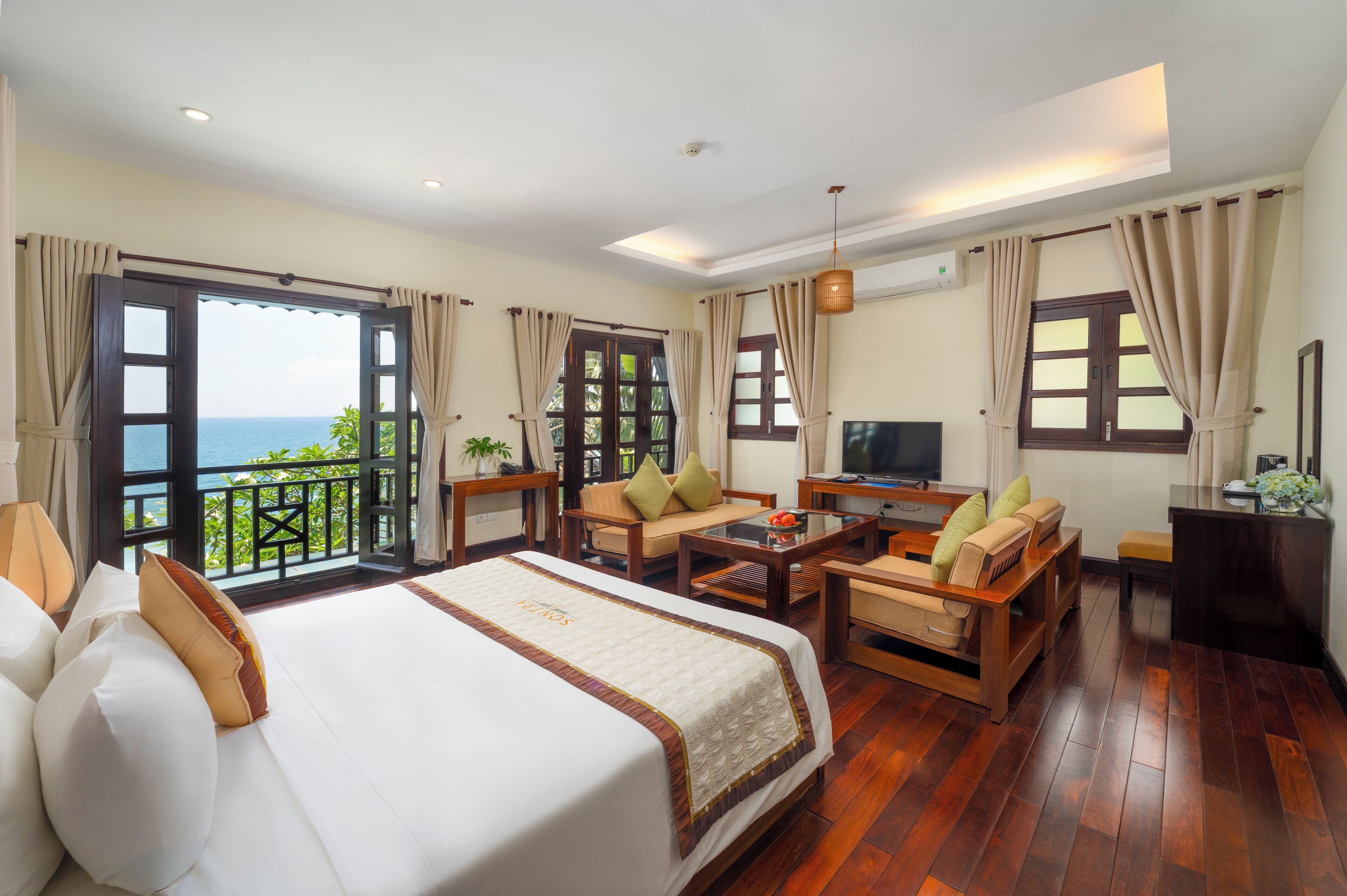 A hotel room with a large bed, a sofa, a TV, and a balcony with a view of the sea.