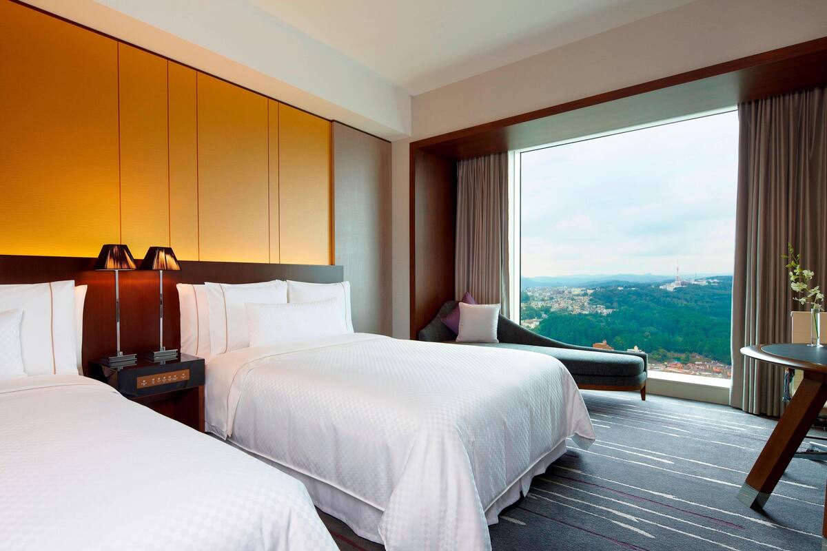 A hotel room with two beds, a nightstand, a view of a cityscape, and a large window with curtains.