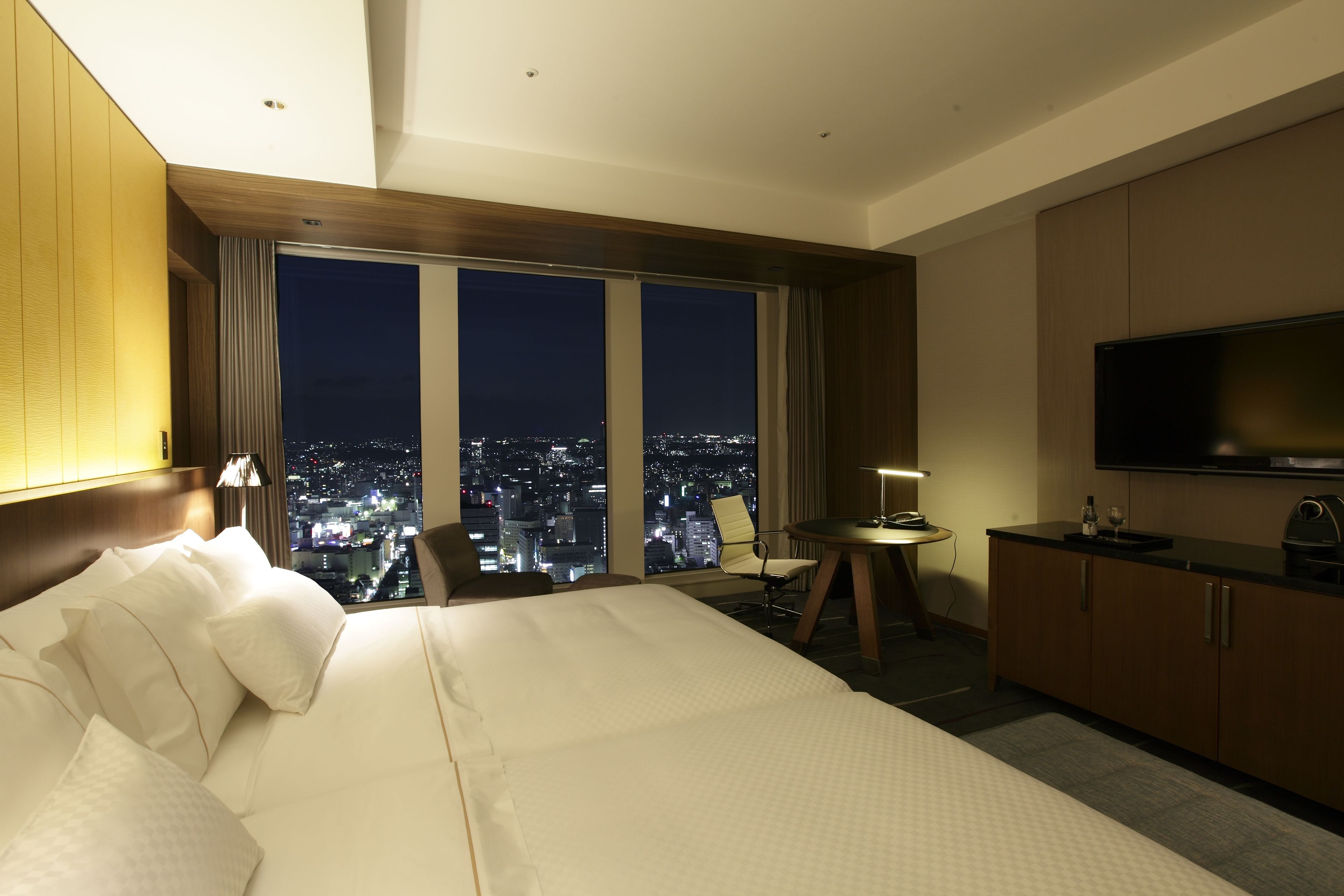 Habitación de hotel con una cama grande, televisión, escritorio con silla y vista a la ciudad de noche.