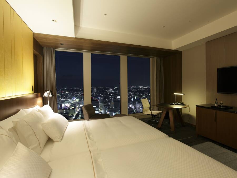 Habitación de hotel con una cama grande, televisión, escritorio con silla y vista a la ciudad de noche.