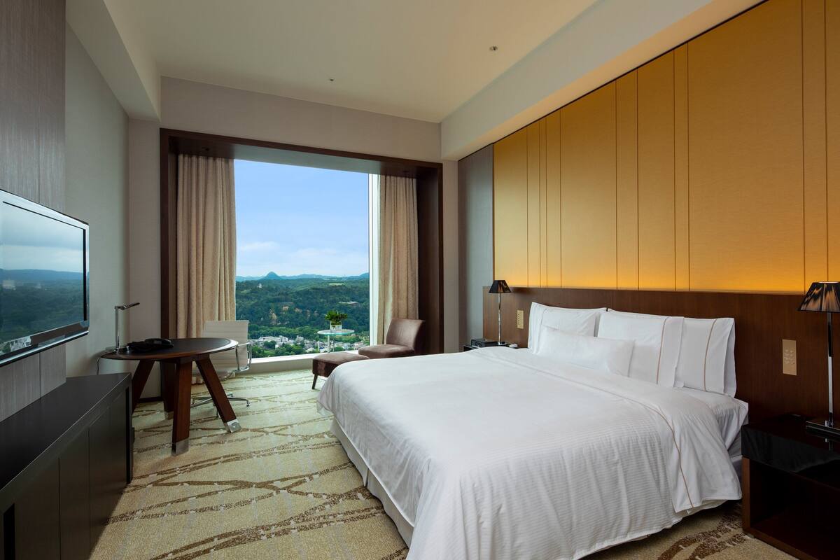 A hotel room with a large bed, a desk with a chair, a TV, a window with curtains, and a view of the mountains.