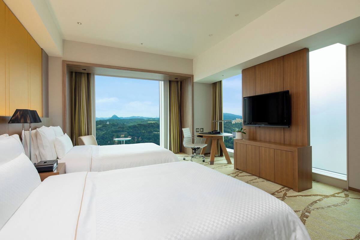 A hotel room with two beds, a TV, a desk, and a view of the mountains.