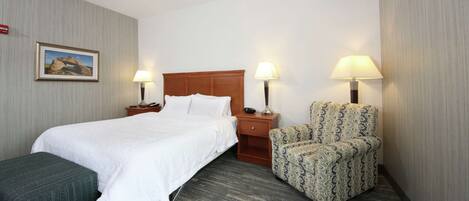 A hotel room with a bed, two bedside tables with lamps, a patterned armchair, a bench, and a framed picture on the wall.