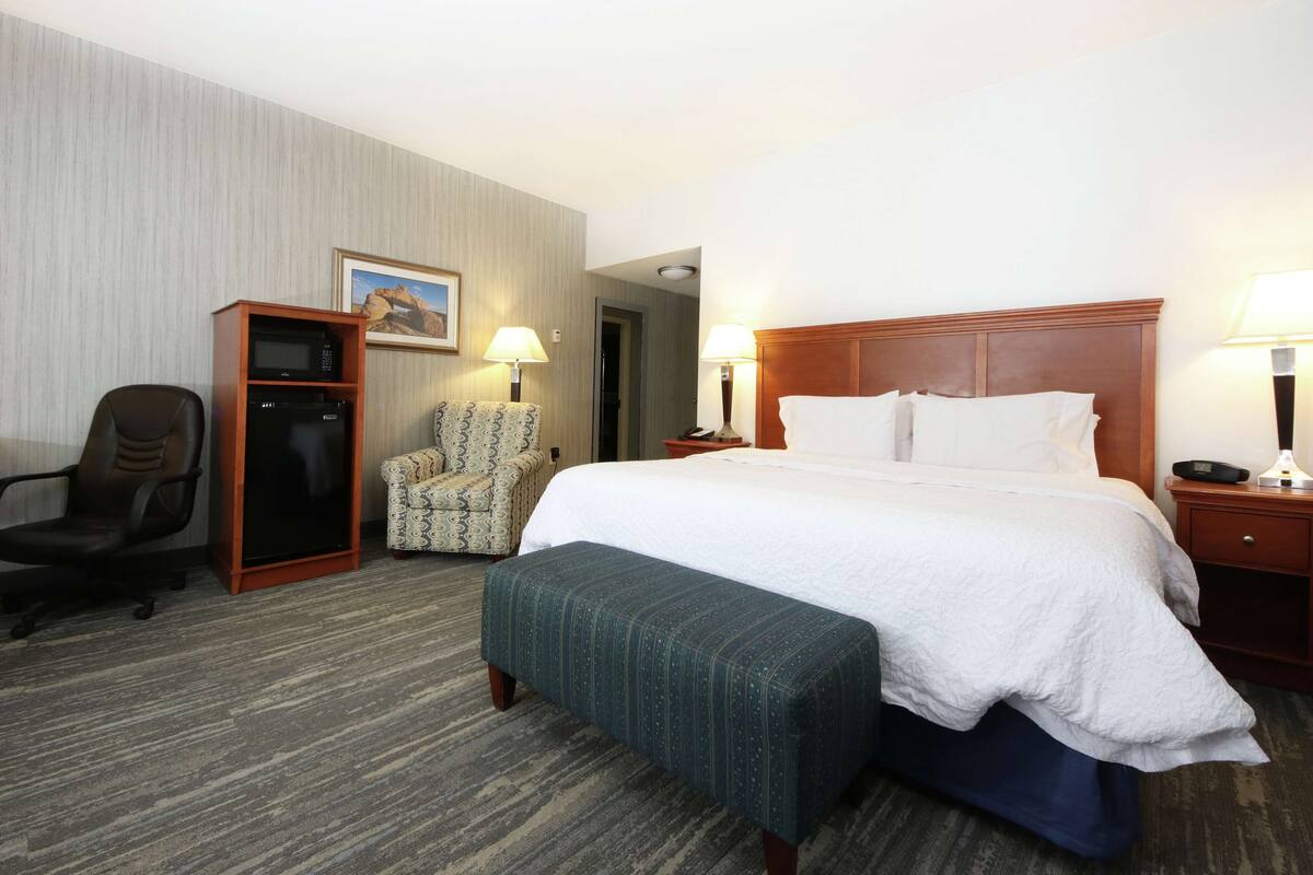 A hotel room with a bed, a desk, a chair, a microwave, and a refrigerator.