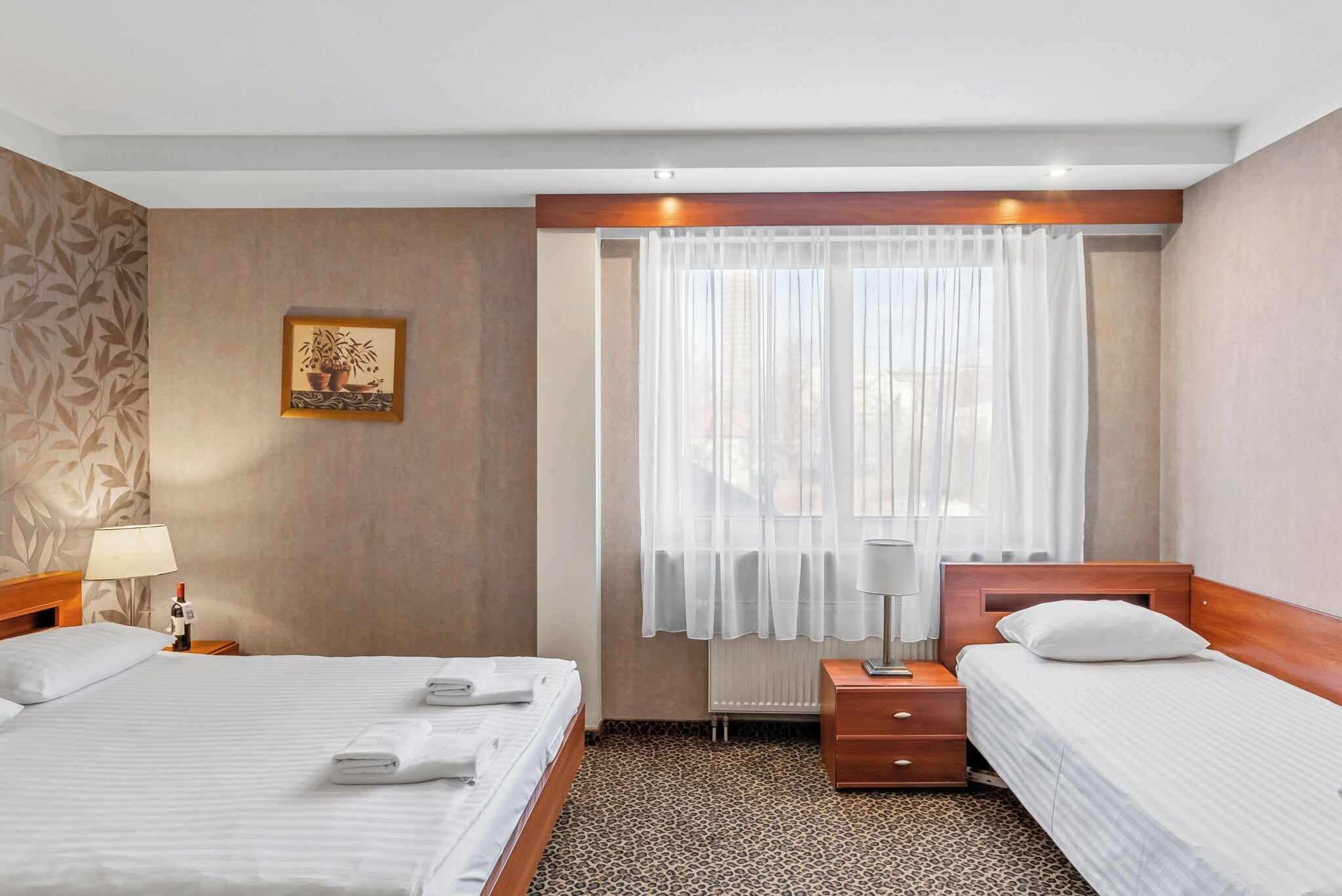Habitación de hotel con dos camas, una mesita de noche con lámpara, ventana con cortinas traslúcidas y alfombra estampada.