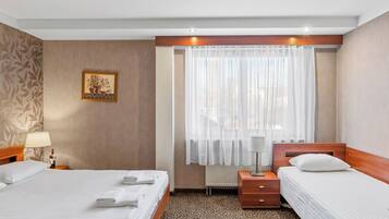Habitación de hotel con dos camas, una mesita de noche con lámpara, ventana con cortinas traslúcidas y alfombra estampada.