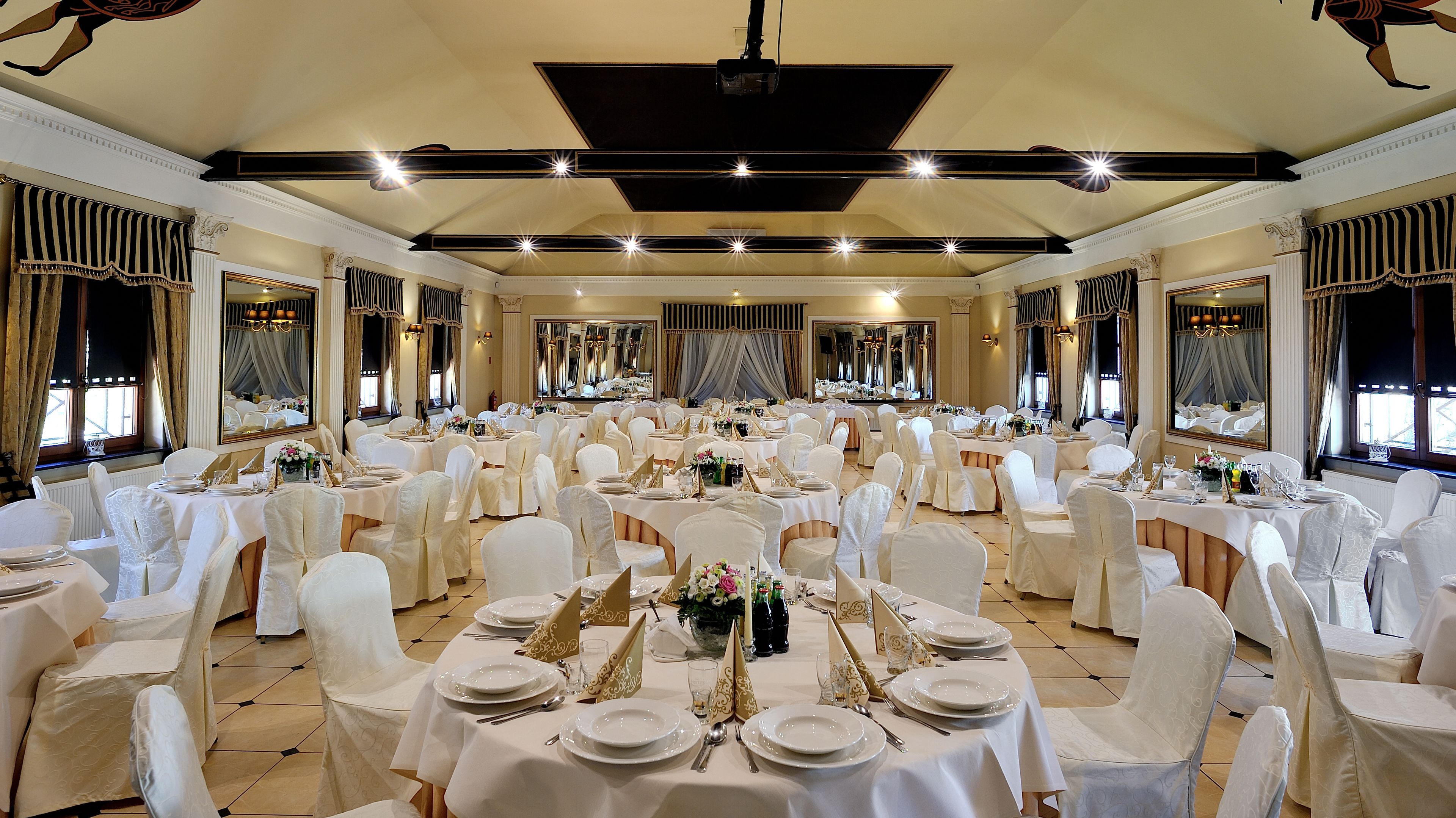 A large banquet hall with round tables set for a formal event, featuring white tablecloths, gold centerpieces, and white chairs.