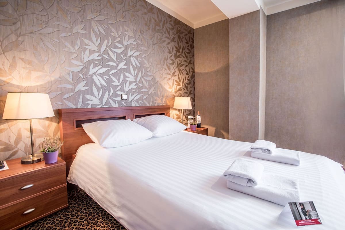 A hotel room with a bed, two bedside tables, a nightstand, a lamp, a potted plant, and a wall with a leaf pattern.