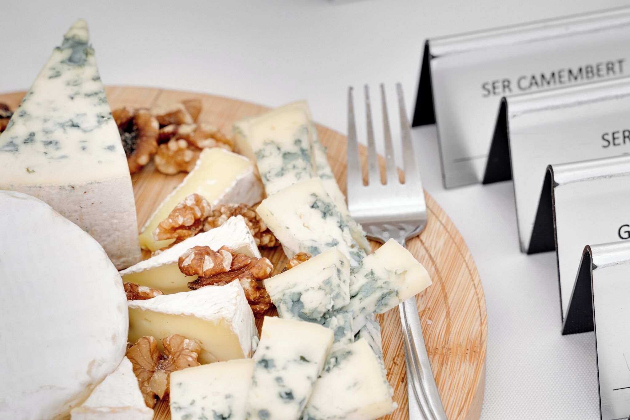 Una tabla de madera con varios quesos y nueces, junto con un tenedor y sujetapapeles etiquetados 'SER CAMEMBERT'.
