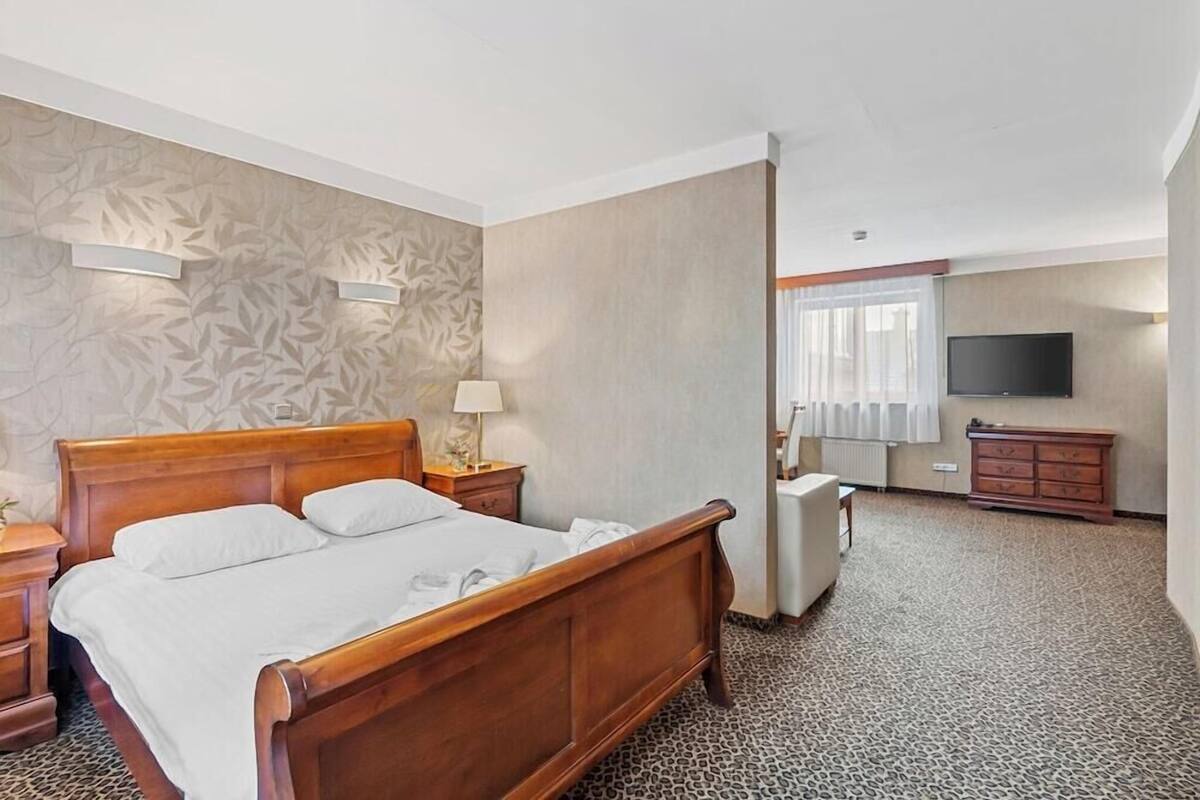 Ein Hotelzimmer mit einem hölzernen Bett, einem Nachttisch, einer Lampe, einer Kommode, einem Fernseher und einem gemusterten Teppich.