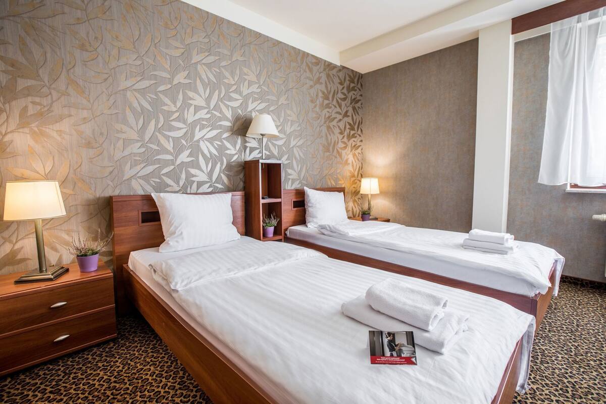 Ein Hotelzimmer mit zwei Betten, einem hölzernen Kopfteil, einem Nachttisch mit Lampe, einer kleinen Ablagefläche mit Pflanze und einem Teppich mit Leopardenmuster.
