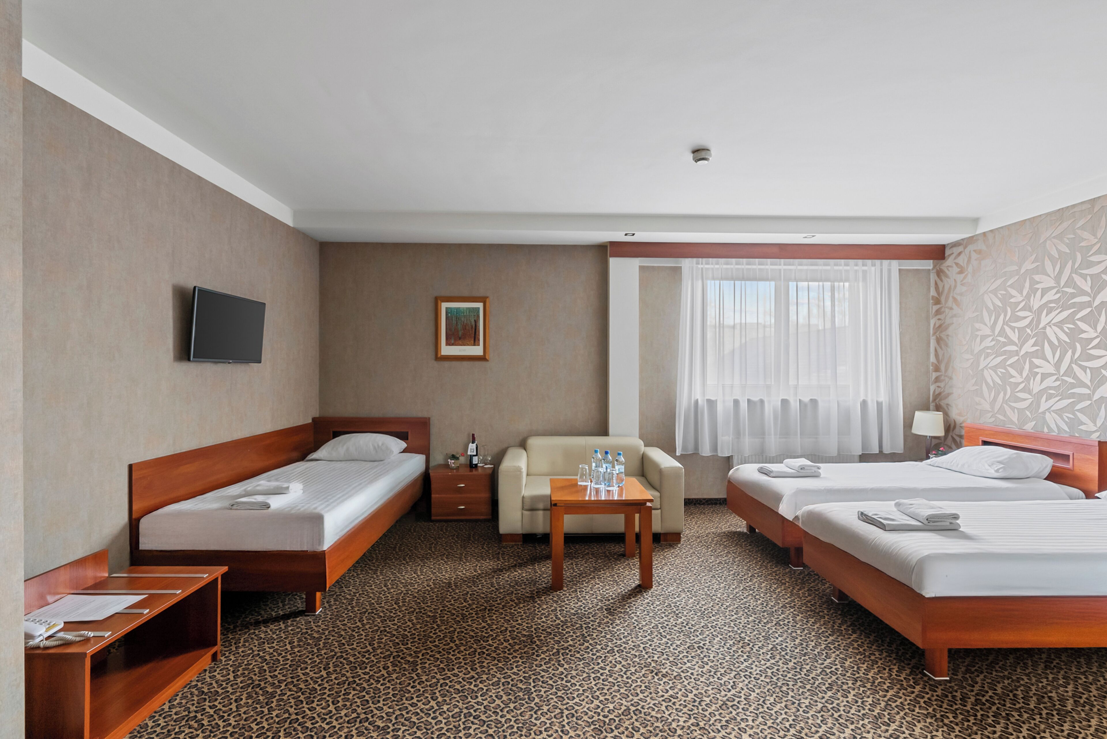 A hotel room with two single beds, a sofa, a TV, a small table with water bottles, and a window with curtains.