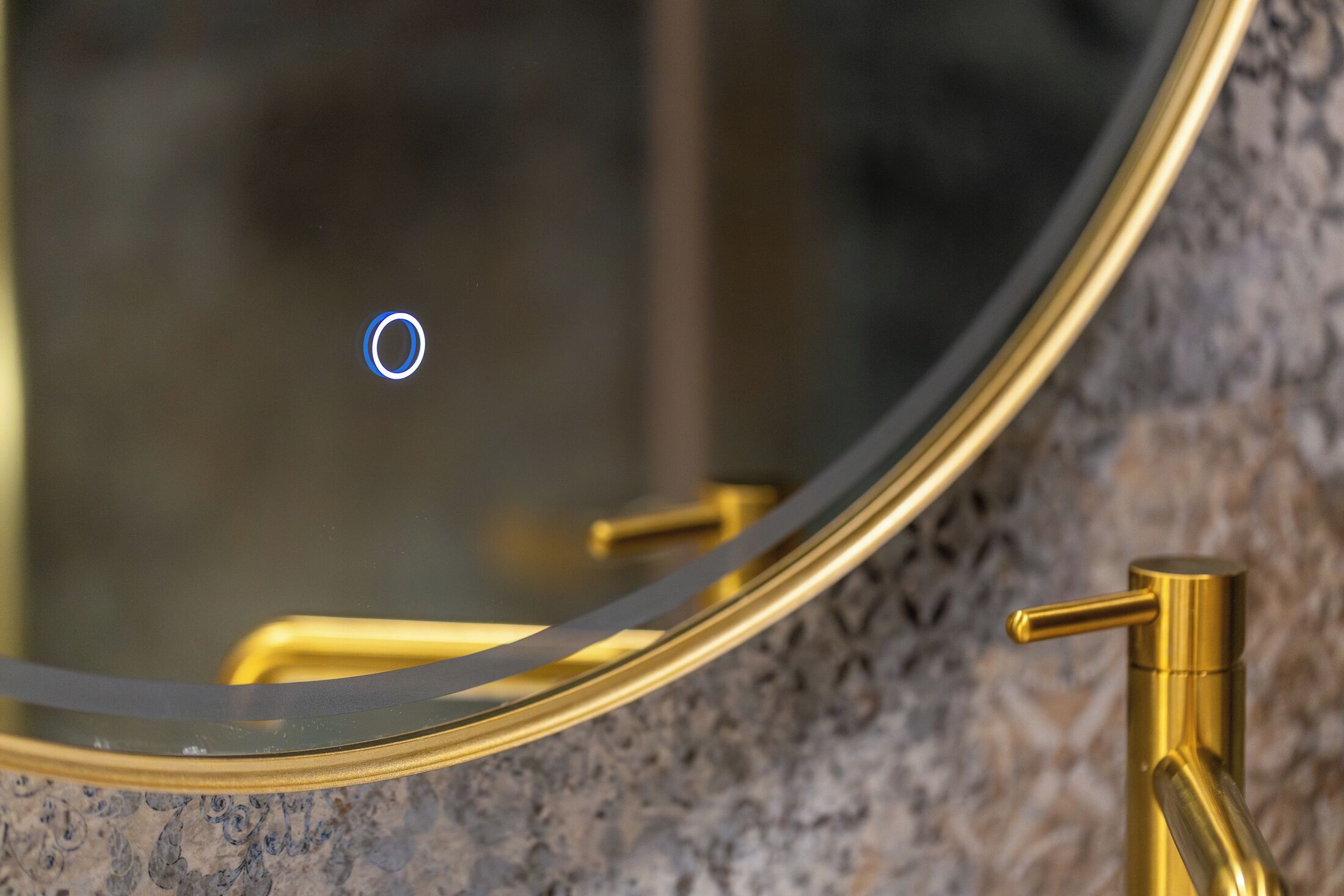 A bathroom with a gold faucet, a round mirror, and a granite countertop.