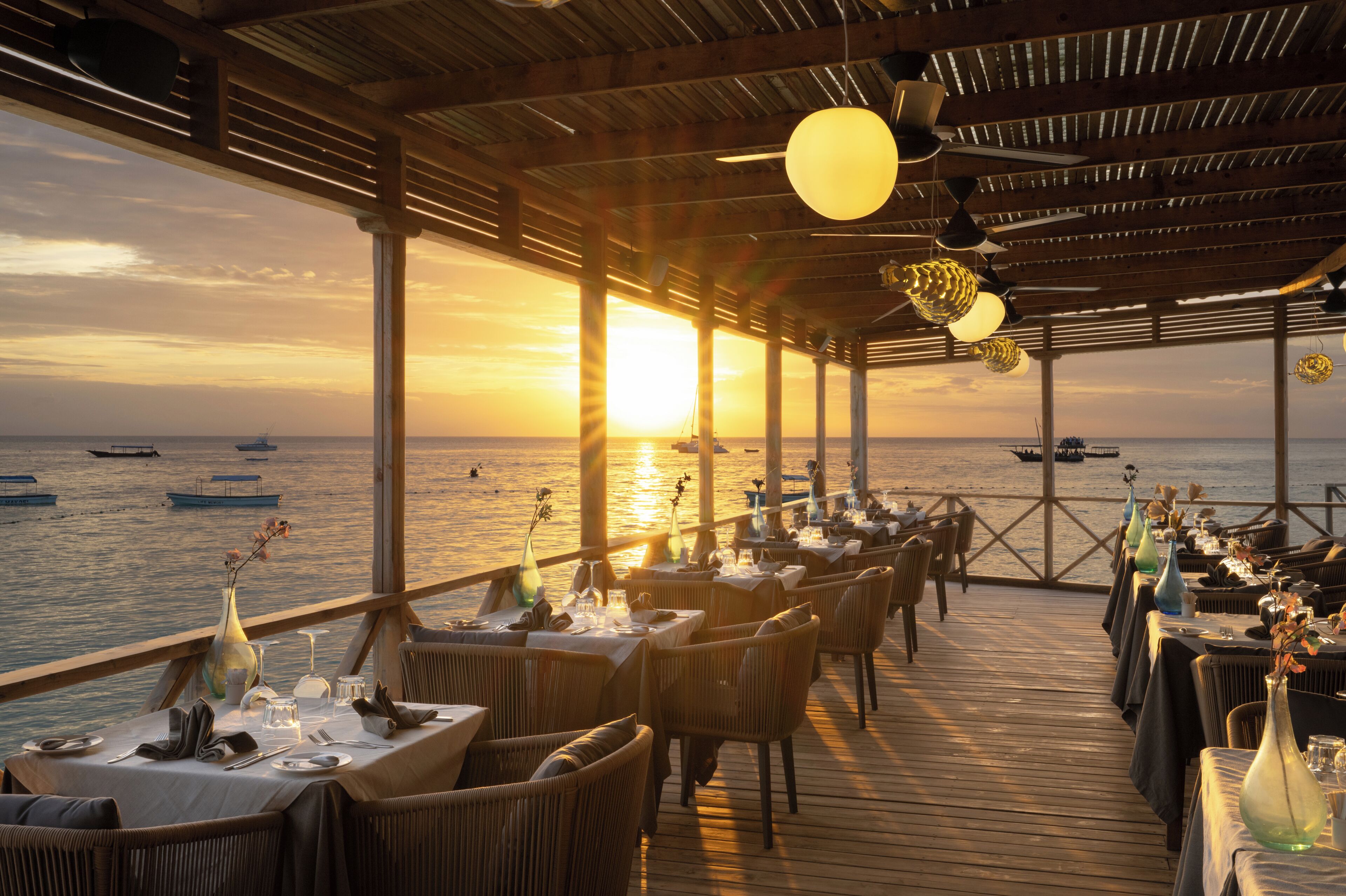 Un restaurant dont les tables sont dressées pour le service, offrant une vue sur la mer et les bateaux qui naviguent au loin.