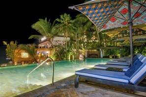 Un espace aménagé au bord de la piscine, avec des chaises longues et des parasols, des palmiers, et un bâtiment sur lequel on peut lire l’inscription « Sixxy Fish ».