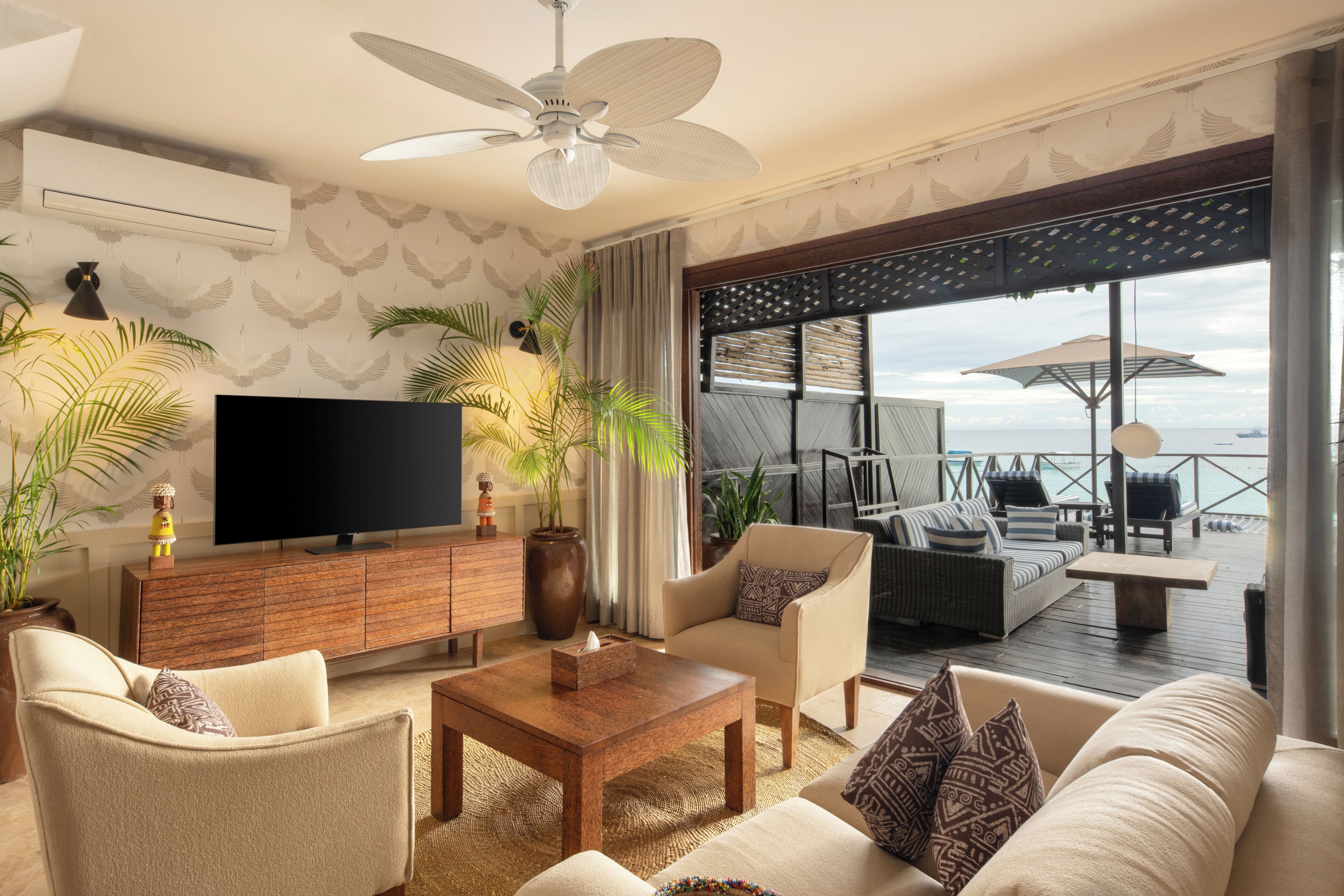A modern living room with a flat-screen TV, comfortable seating, a ceiling fan, and a view of the sea through large windows.