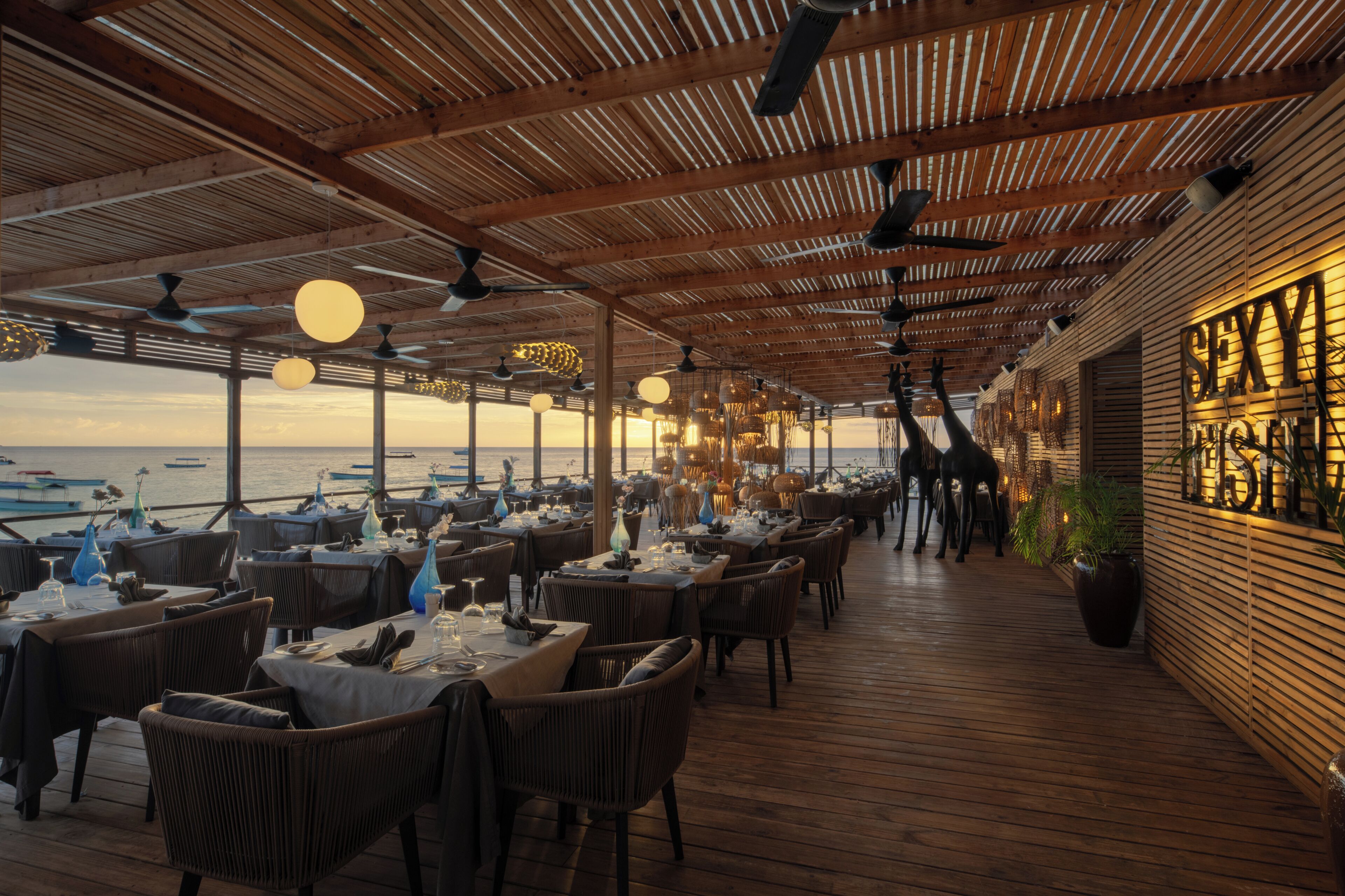 En udendørs restaurant med trægulv og borde dækket til spisning, med udsigt over vand.