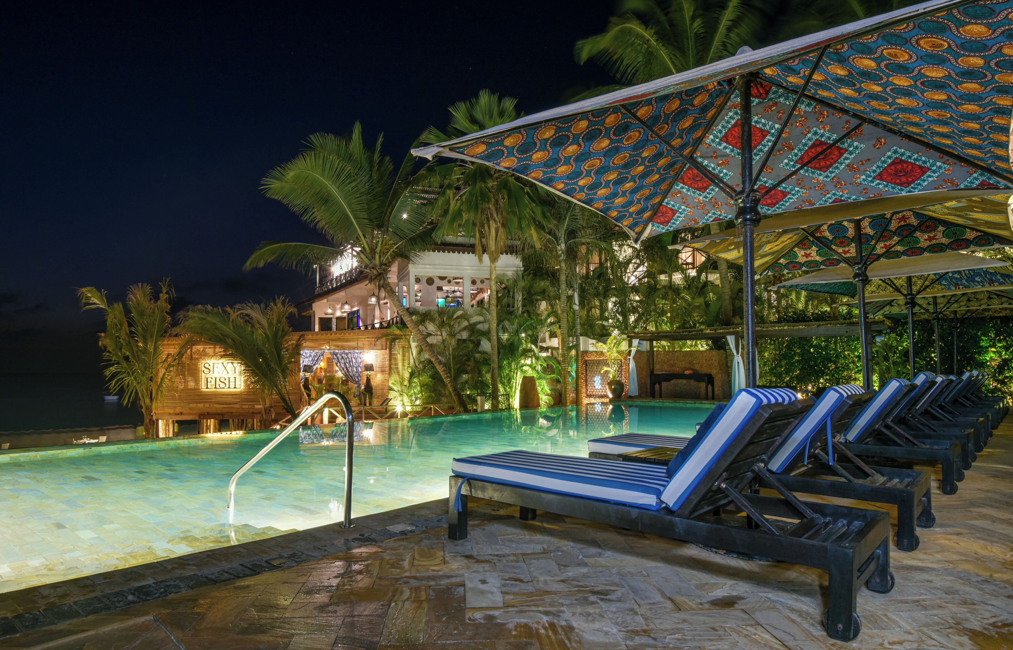 Un espace piscine avec des chaises longues et des parasols, un palmier et un bâtiment sur lequel on peut lire l’inscription « SEXY FISH ».