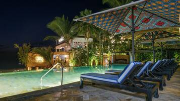 Un espace piscine avec des chaises longues et des parasols, un palmier et un bâtiment sur lequel on peut lire l’inscription « SEXY FISH ».