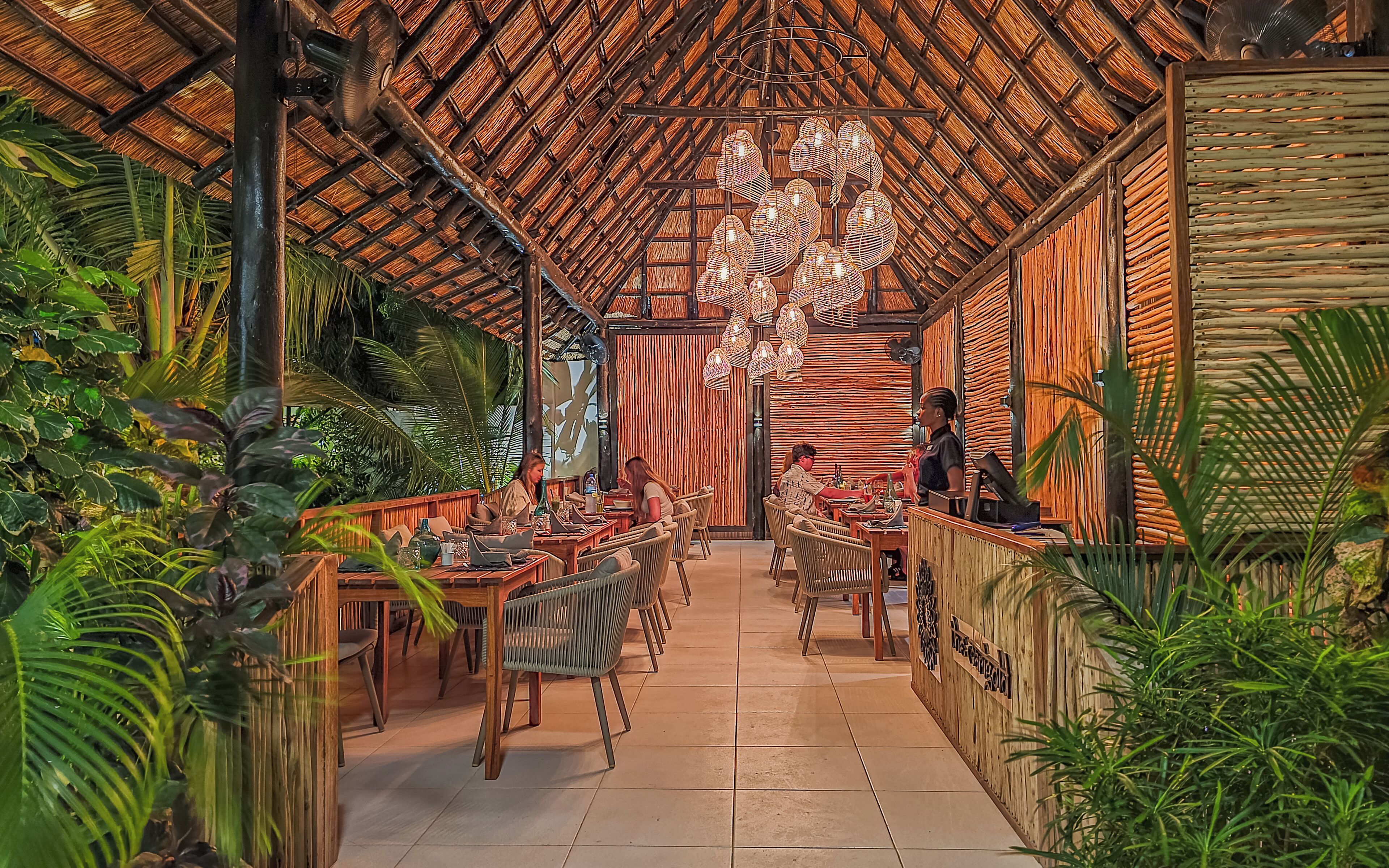 An indoor restaurant with a thatched roof, wicker furniture, and tropical plants.