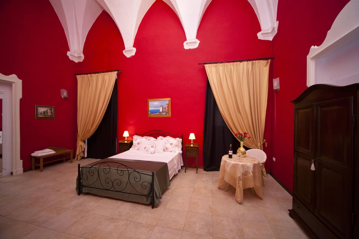 A red-walled bedroom with a bed, two bedside tables with lamps, a small table with a bottle and flowers, a bench, and a wardrobe.
