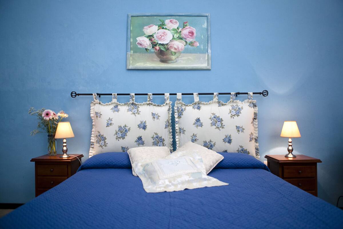 A bedroom with a blue bedspread, floral-patterned headboard, two bedside tables with lamps, and a framed floral painting on the wall.