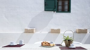 A table with breakfast items, a potted plant, and wicker baskets against a white wall with a green window.