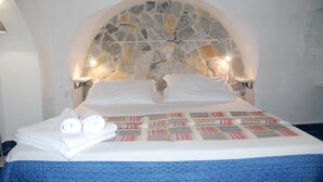 A neatly made bed with white linens, a patterned bedspread, and two rolled towels on the side.