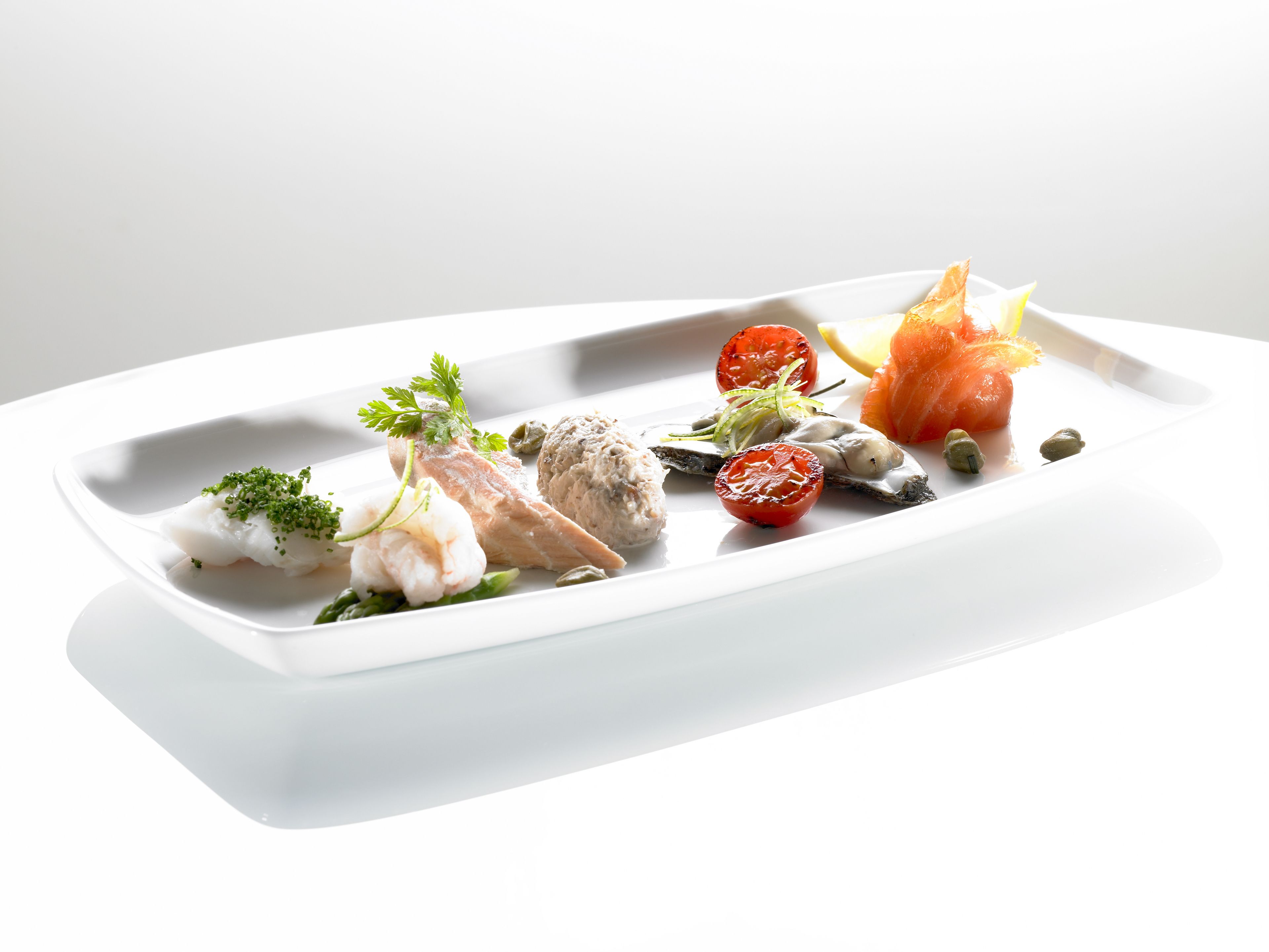 Een wit, rechthoekig bord met diverse zeevruchten, waaronder vis, garnalen en een stukje zalm, versierd met kruiden.