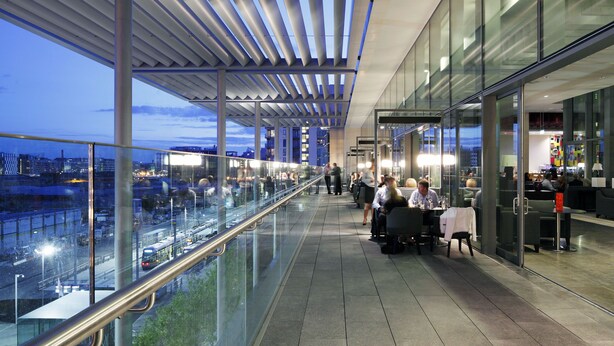 Et moderne udendørsområde med glasgelænder og udsigt over en by, med et overdækket siddeområde og en glasfacade.