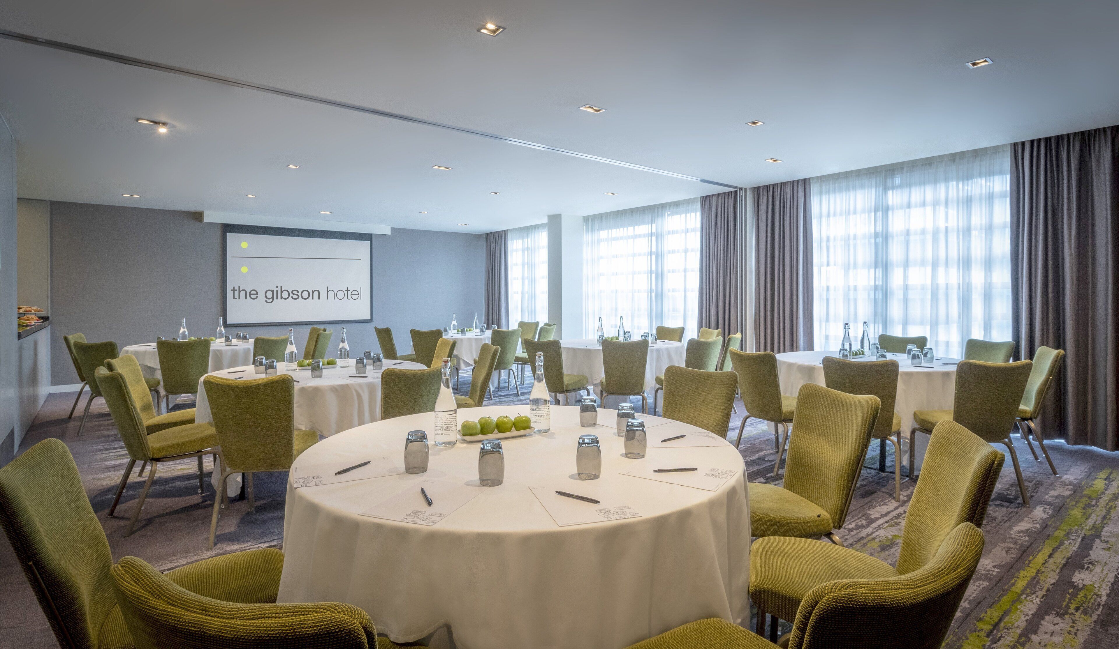 Un salón de conferencias con mesas redondas, sillas y una pantalla de proyector que muestra 'the gibson hotel'.