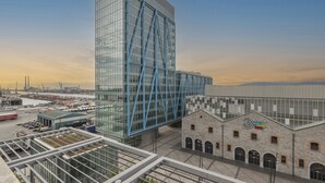 En moderne bygning med en glasfacade og blålig konstruktion, beliggende ved siden af en historisk bygning i sten med et skilt, der siger "Arena".