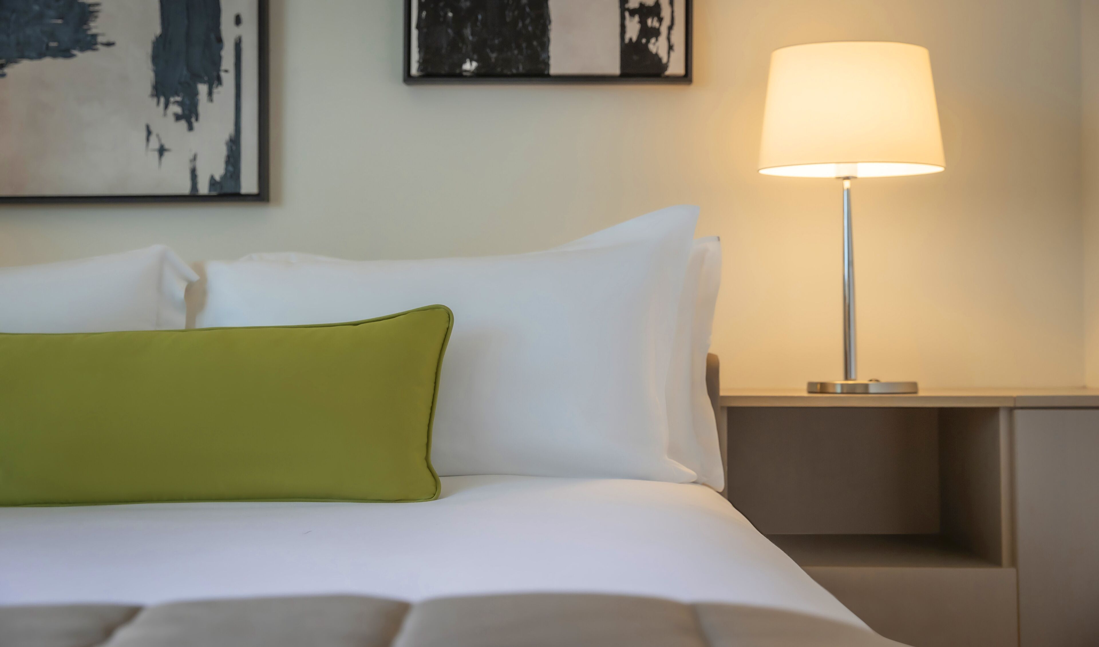 A neatly made bed with white and green pillows, a bedside lamp, and abstract art on the wall.