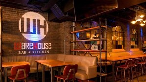 Een restaurantinterieur met een bordje 'Warehouse Bar + Kitchen', houten tafels, rode stoelen en een beige zithoek.