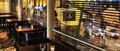Un restaurante con mesas y sillas, vistas a la calle y una barandilla con listones horizontales.