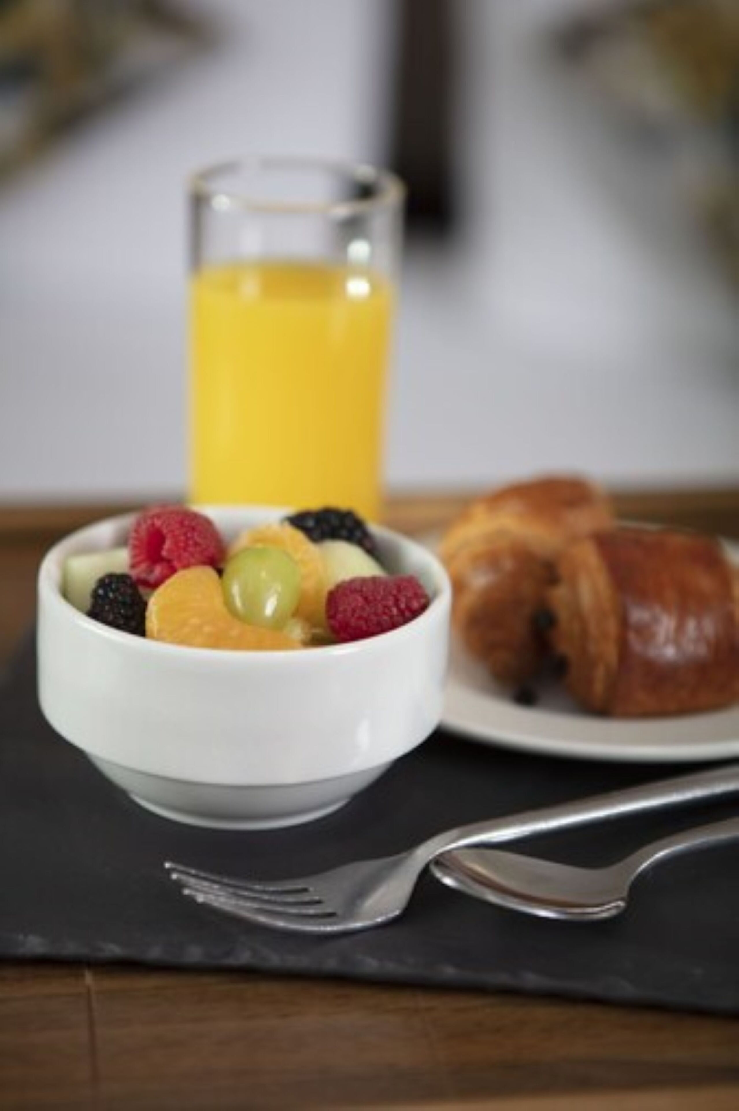 Eine Schale mit Obst und ein Croissant auf einem Teller, im Hintergrund ein Glas Orangensaft.