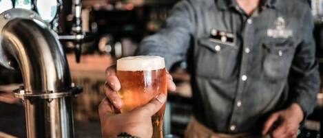 Una persona sosteniendo una cerveza en un bar, con una grifo y un cantinero al fondo.