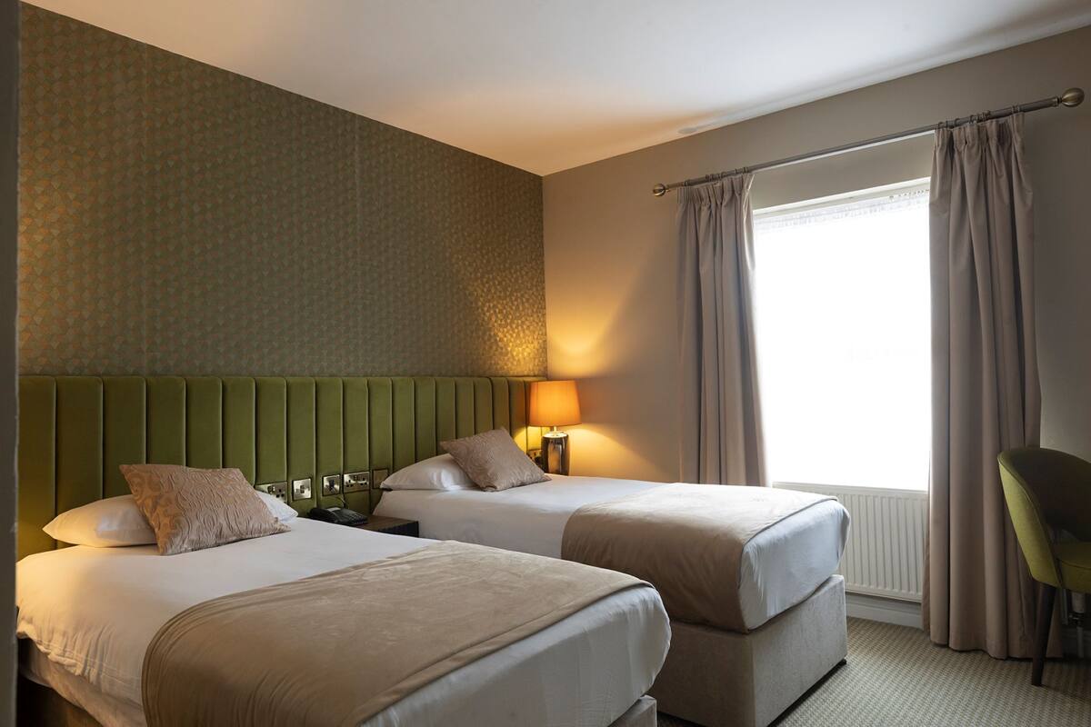 A hotel room with two beds, a green headboard, a window with curtains, and a chair.