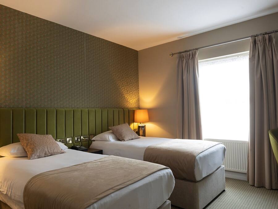 Ein Hotelzimmer mit zwei Betten, einem grünen Kopfteil, einem Fenster mit Vorhängen und einem Sessel.