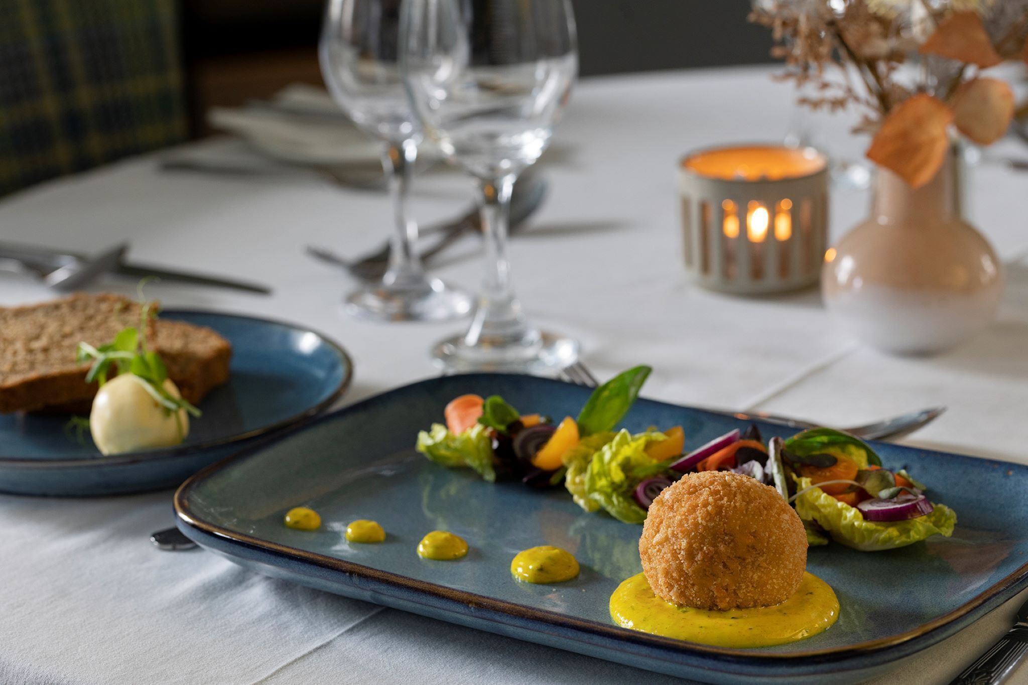 Ein Esstisch mit einem blauen Teller, der einen Salat, ein Brötchen und ein kleines, rundes Objekt, möglicherweise eine Croquette, mit gelber Soße enthält.