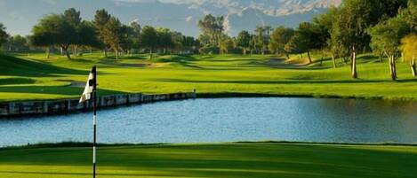 A golf course with a water hazard, trees, and mountains in the background.
