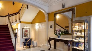 A grand staircase with a red carpet, a blue armchair, a display cabinet with framed pictures, and a floral arrangement on a console table. - Cahir House Hotel (Cahir)