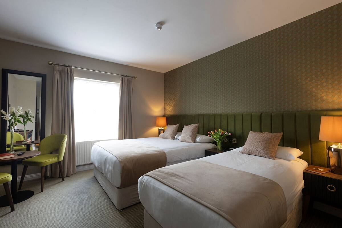 A hotel room with two beds, a desk with a chair, a mirror, and a window with curtains.